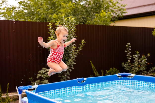 Gelukkig klein meisje in rode zwembroek springen in het buitenzwembad thuis. Babymeisje die leren zwemmen. Waterpret voor kinderen.