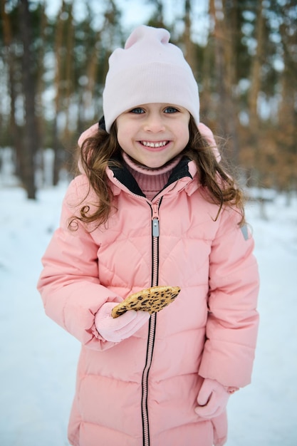 Gelukkig klein meisje in roze pastel warme winterkleren houdt koekjes in haar hand en glimlacht met een brede glimlach kijkt naar de camera tijdens het spelen in een besneeuwd bos Geniet van de prachtige winter buiten