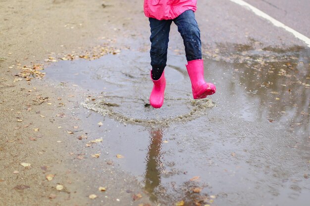 Gelukkig klein meisje in roze waterdichte jas rubberen laarzen springt vrolijk door plassen op straat weg bij regenachtig weer Lente herfst Kinderplezier in de frisse lucht na regen Openlucht recreatie