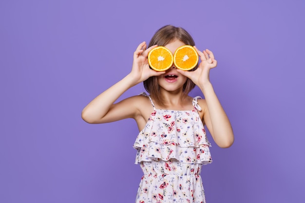 Gelukkig klein meisje in zomerjurk en strohoed met een sinaasappel op paarse achtergrond