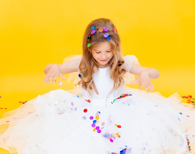 Gelukkig klein meisje met blond haar en in een witte jurk vangt confetti op een gele achtergrond, vakantieconcept