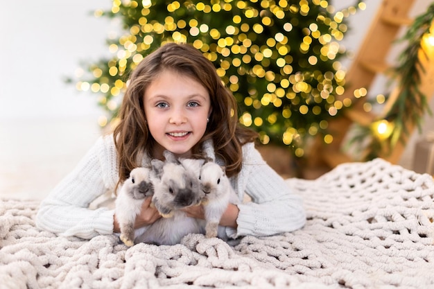 Foto gelukkig klein meisje met drie konijnen symbolen van het nieuwe jaar 2022 omarmt dieren op de achtergrond van een kerstboom met wazig licht