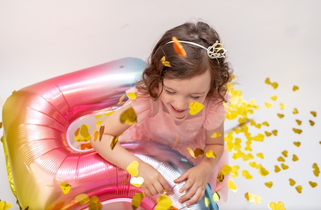 Gelukkig klein meisje met een ballon nummer vijf speelt met confetti op een witte feestelijke achtergrond. vakantie verjaardag