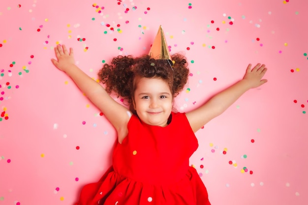 Gelukkig klein meisje met een verjaardagsmuts ligt tussen de veelkleurige confetti op een roze achtergrond, een uitzicht van bovenaf