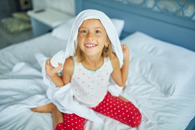 Foto gelukkig klein meisje wordt wakker, vrolijk, deken op het hoofd, uit slaap op een groot en gezellig bed wit linnen thuis