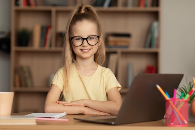 Gelukkig klein schoolmeisje dat een laptop gebruikt die een bril draagt om thuis te zitten