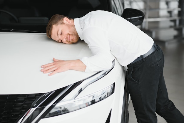 Gelukkig knappe bebaarde man die een auto koopt bij een dealer die de motorkap van een nieuwe auto knuffelt