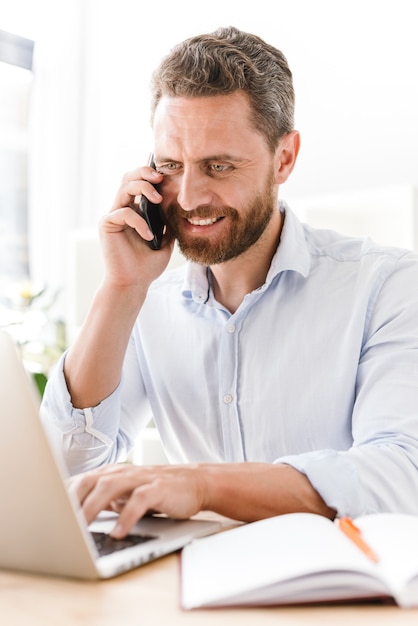 Gelukkig knappe bebaarde man praten via de mobiele telefoon tijdens het gebruik van laptopcomputer.