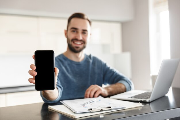 Gelukkig knappe man freelancer zittend aan de keukentafel, werken op laptopcomputer, leeg scherm mobiele telefoon tonen