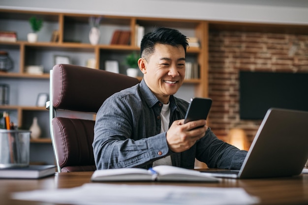 Gelukkig knappe volwassen Aziatische zakenman typen op smartphone op werkplek op laptop in huis