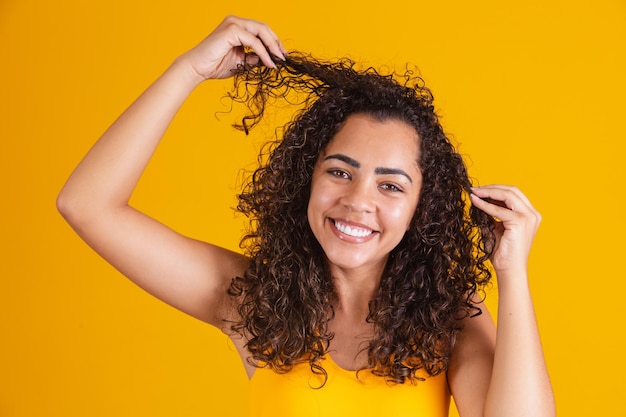 Gelukkig lachen Afro-Amerikaanse vrouw met haar krullend haar op gele achtergrond. Lachende krullende vrouw in gele outfit die haar haar aanraakt en naar de camera kijkt.