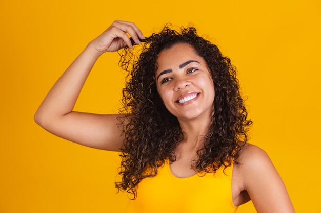 Gelukkig lachen afro-amerikaanse vrouw met haar krullend haar op gele achtergrond. lachende krullende vrouw in gele outfit die haar haar aanraakt en naar de camera kijkt.