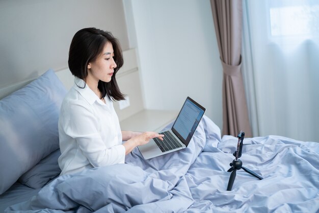 Gelukkig lachend Aziatische jonge vrouw met laptopcomputer en mobiele telefoon met video-oproep in de slaapkamer