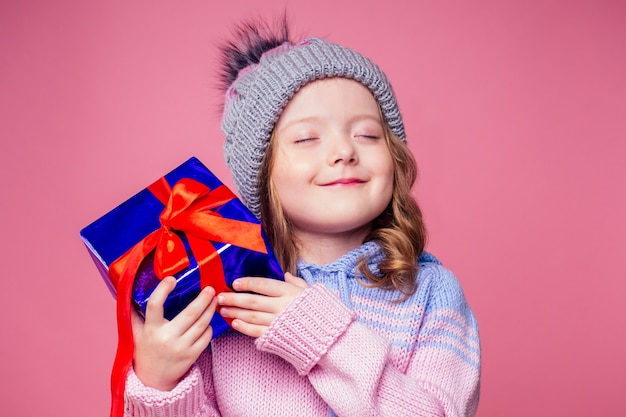 Gelukkig lachend blond krullend kapselmeisje in gebreide grijze muts met kerstcadeaudoos met strik op roze achtergrondstudio. nieuwjaarscadeau in handen van een vrouwelijk kind dat een wens copyspace maakt.