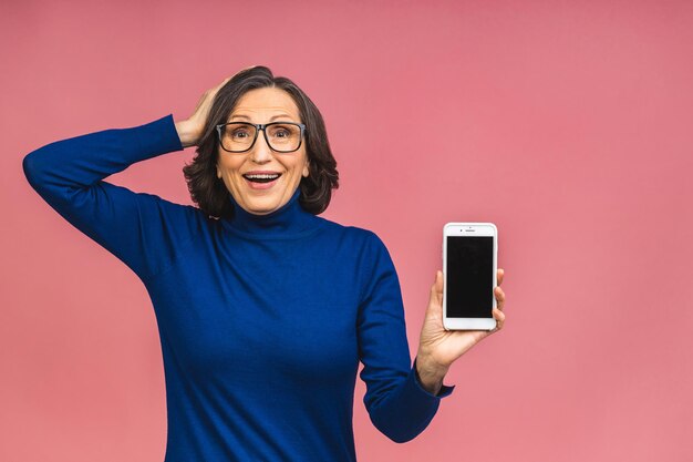 Gelukkig lachend gelukkig senior volwassen vrouw in casual met lege smartphone scherm terwijl kijken naar de camera geïsoleerd over roze achtergrond. Telefoon gebruiken.