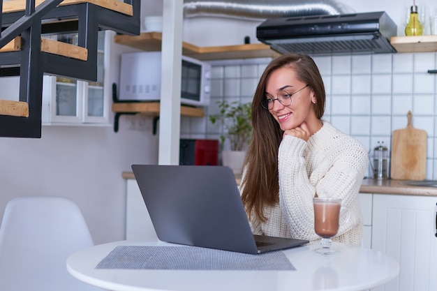 Gelukkig lachend gezellige vrouw dragen een warme witte trui en een ronde bril met een kopje warme chocolademelk kijken naar online video op laptop op comfortabele huiselijke keuken