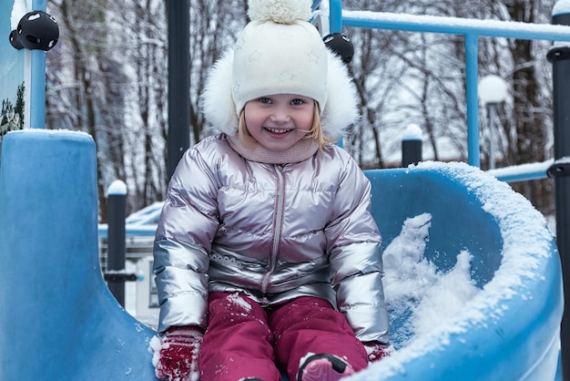 Gelukkig lachend klein meisje rijdt glijbaan in bevroren winter besneeuwde speelplaats in warme kleding Schattig kind in heldere winterkleren