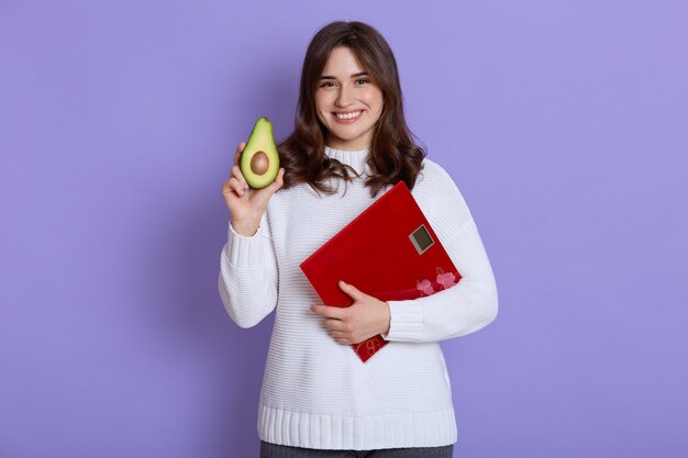 Gelukkig lachend mager wijfje dat witte sweater draagt die vloerschalen en de helft van avocado in handen houdt