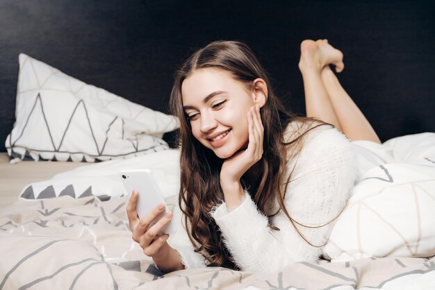 Gelukkig lachend meisje in pyjama liggend op bed op zoek naar haar smartphone goed humeur