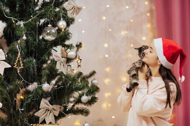 Gelukkig lachend meisje in rode kerstman hoed houdt kleine kitten. Kerstboom is versierd met sprankelende zilveren ballen en guirlandes.