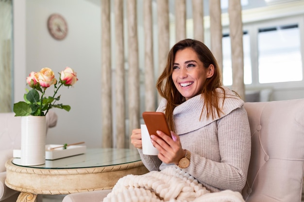 Gelukkig lachend mooi meisje met smartphone-apparaat in gezellig huis, aantrekkelijke jonge vrouw chatten met vrienden op sociaal netwerk terwijl zittend op de bank thuis en koffie drinken