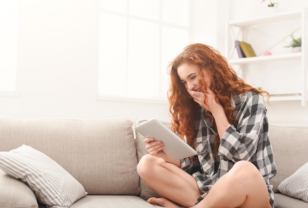 Gelukkig lachend roodharig meisje dat op digitale tablet leest, zittend op een beige bank in vrijetijdskleding, kopieer ruimte