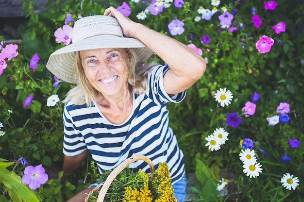 Foto gelukkig lachend senior vrouw poseren in zomertuin met bloemen in strooien hoed.