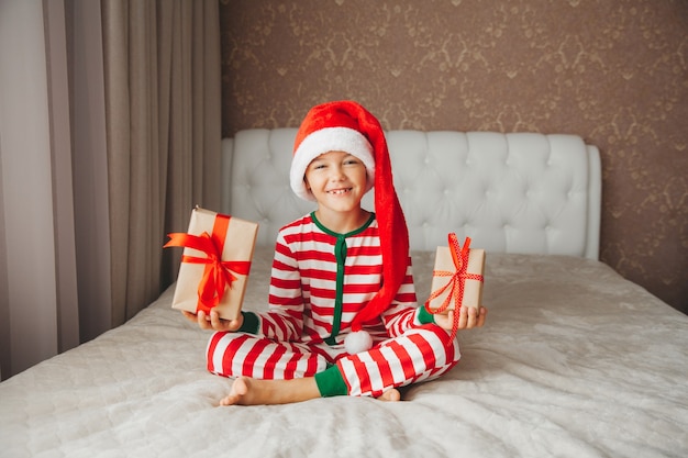 Gelukkig lachend vrolijk kind jongen in kerstman hoed, met een kerstcadeau in zijn handen, zittend op het bed.