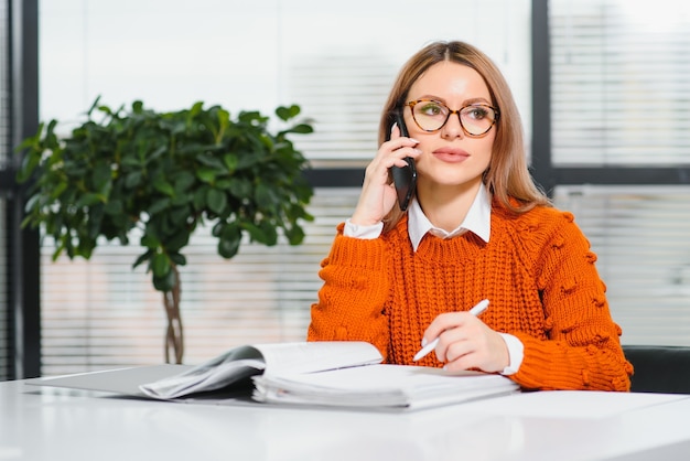 Gelukkig lachend zakenvrouw op het werk praten over de telefoon, zittend op haar werkplek in kantoor, kopieer ruimte