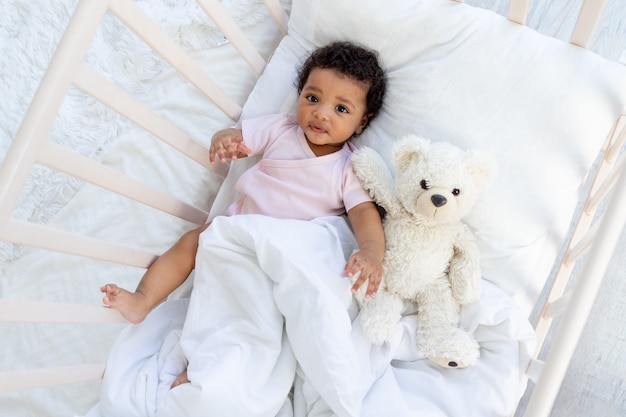 Gelukkig lachende afro-amerikaanse baby in een wieg met een teddybeer valt in slaap of gaat naar bed