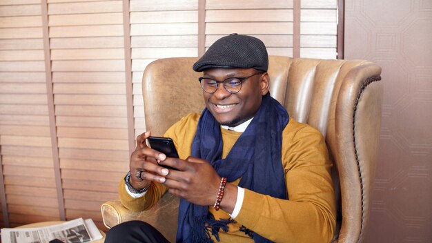 Gelukkig, lachende Afro-Amerikaanse man zittend op een stoel in een chique dure restaurant, café met een smartphone, telefoon in de hand
