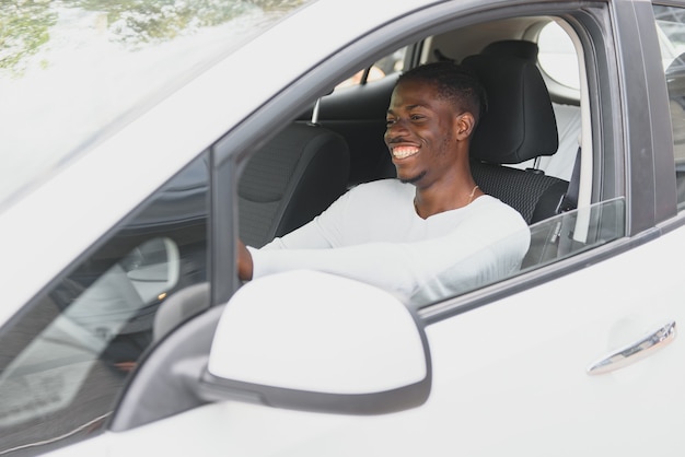 Gelukkig lachende Afro-Amerikaanse mannelijke bestuurder zit achter het zelfrijdende stuur van een autonome elektrische moderne auto. Gelukkige kerel houdt telefoon vast en glimlacht naar camera in moderne elektrische auto