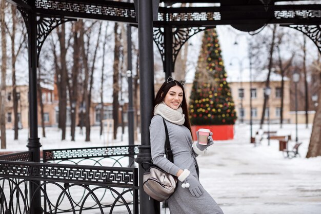 Gelukkig lachende brunette vrouw rusten in winter park, koffie drinken, staande voor de kerstboom, buiten in de winter.