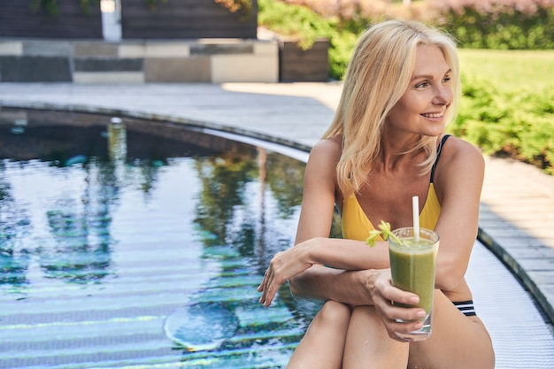 Gelukkig lachende dame met een glas smoothie in de hand wegkijkend terwijl ze bij het zwembad zit