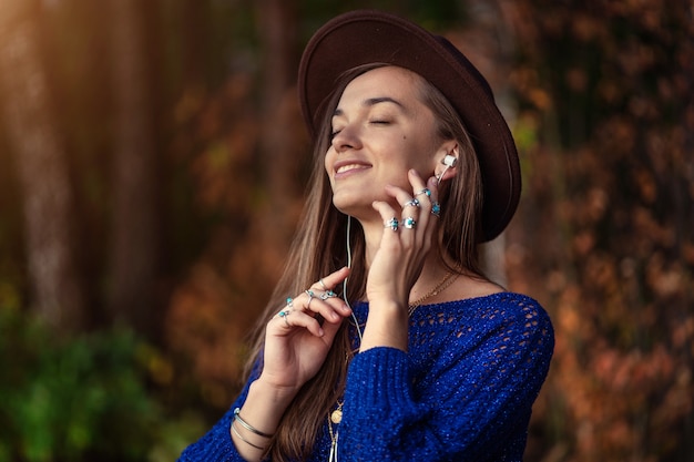 Gelukkig lachende herfst brunette vrouw in een bruine hoed en in een gebreide trui met zilveren ringen met turquoise steen geniet van het luisteren naar herfstmuziek op koptelefoon buitenshuis in de herfst