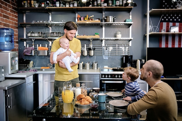 Gelukkig lachende homoseksuele man die pasgeboren baby voorstelt aan zijn oudste zoon bij het familieontbijt