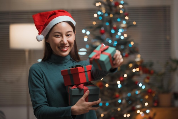 Gelukkig lachende jonge Aziatische vrouw in kerstmuts met veel geschenkdoos met kerstboom thuis