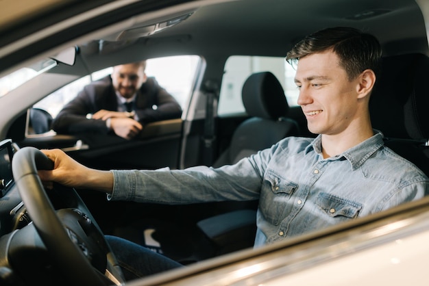 Gelukkig lachende jonge man achter het stuur van nieuwe auto in autodealer. Professionele autoverkoper die een pak draagt, vertelt over een nieuw automodel. Concept van het kiezen en kopen van een nieuwe auto in de showroom.