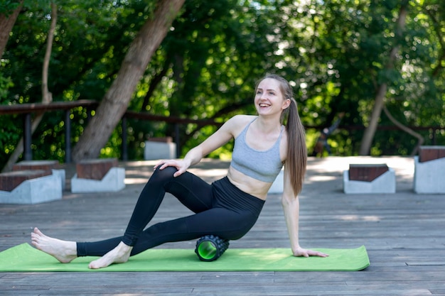 Gelukkig lachende jonge slanke vrouw kneedt de spieren van de dijen met myofasciale roller buitenshuis. Myofasciale vrijlating.