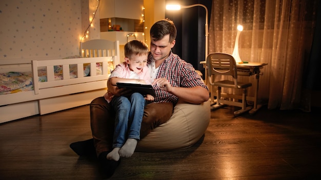 Gelukkig lachende jongen met jonge vader die 's nachts in de slaapkamer zit en spelletjes speelt op tabletcomputer. Concept van kinderopvoeding en familie die 's nachts samen tijd hebben.