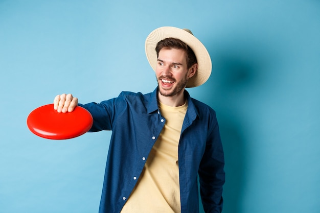 Gelukkig lachende man frisbee vangen tijdens het spelen met vrienden op zomervakantie, staande in strooien hoed op blauwe achtergrond.