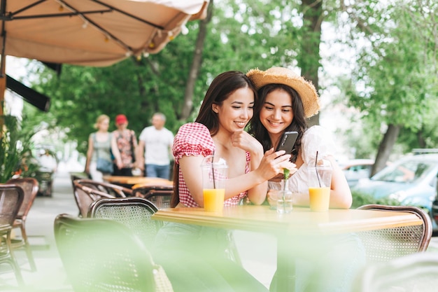Gelukkig lachende mooie brunette jonge vrouwen vrienden in zomerkleren met behulp van mobiele telefoon in café