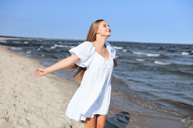 Foto gelukkig lachende mooie vrouw is op het oceaanstrand in een witte zomerjurk met open armen