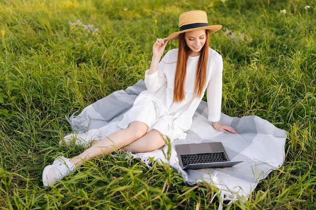 Gelukkig lachende roodharige jonge vrouw met strohoed en witte jurk op zoek naar laptop scherm zittend op prachtige weide van groen gras