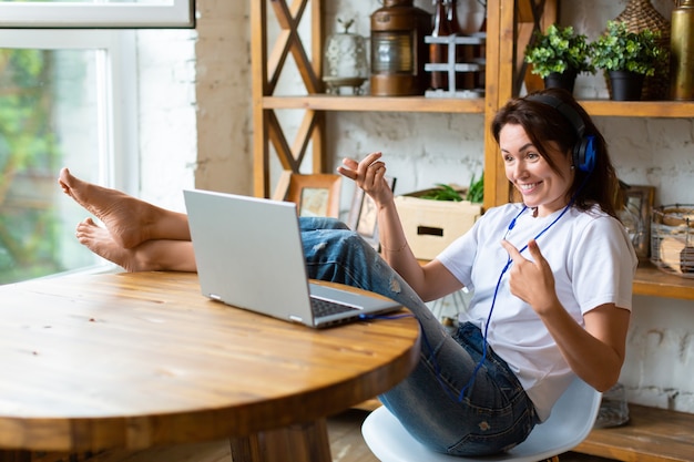 Gelukkig lachende vrouw in blauwe koptelefoon voor een laptop monitor thuis zet ze haar voeten op de...