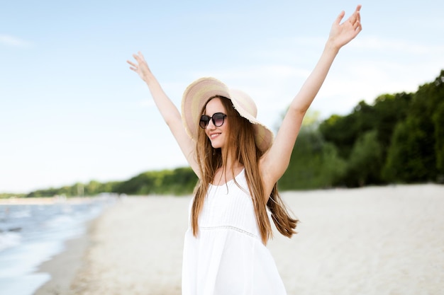 Gelukkig lachende vrouw in vrij geluk gelukzaligheid op oceaan strand staande met een hoed, zonnebril en raspende handen. Portret van een multicultureel vrouwelijk model in witte zomerjurk genietend van de natuur tijdens trav