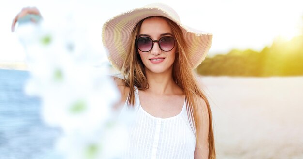 Gelukkig lachende vrouw in vrij geluk gelukzaligheid op oceaan strand staande met hoed zonnebril en witte bloemen Portret van een multicultureel vrouwelijk model in witte zomerjurk genieten van de natuur