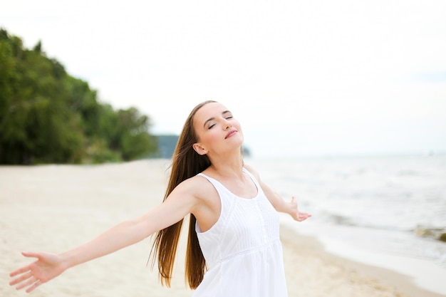 Gelukkig lachende vrouw in vrije gelukzaligheid op oceaanstrand staande met open handen. Portret van een multicultureel vrouwelijk model in witte zomerjurk, genietend van de natuur tijdens reisvakanties