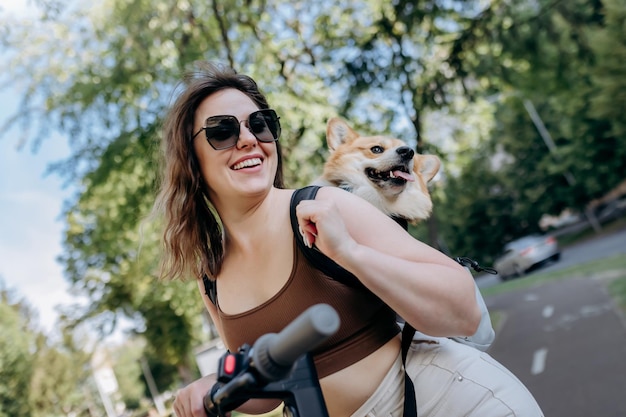 Gelukkig lachende vrouw reiziger rijdt op haar elektrische scooter in stadspark met hond Welsh Corgi Pembroke in een speciale rugzak