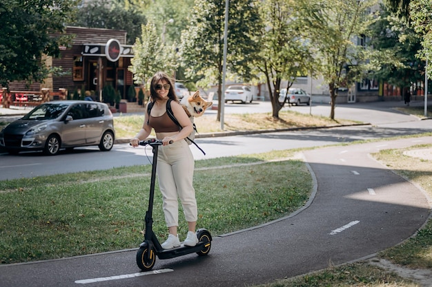 Gelukkig lachende vrouw reiziger rijdt op haar elektrische scooter in stadspark met hond Welsh Corgi Pembroke in een speciale rugzak